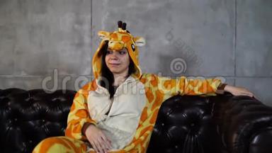 穿长颈鹿服装的女人坐在沙发上。 穿着长颈鹿滑稽睡衣，坐在皮榻上微笑的年轻女子